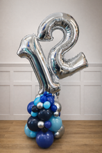 Load image into Gallery viewer, Silver number 12 balloon with blue and silver cluster on a wooden floor against a white paneled wall.