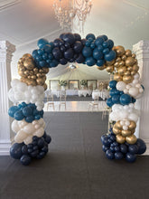 Load image into Gallery viewer, A large balloon arch decoration in navy blue, metallic gold, and white, set up in a decorated event hall with tables and chairs visible in the background.
