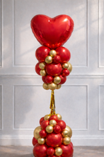 Load image into Gallery viewer, Balloon sculpture with red heart and red and gold balloons against a white paneled wall.