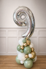 Load image into Gallery viewer, Silver number 9 balloon with green, gold, and white balloons against a white paneled wall.
