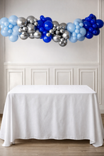 Load image into Gallery viewer, Balloon arch with blue, silver, and gray balloons above a white table.

