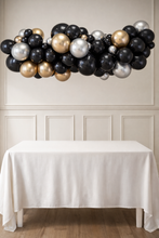 Load image into Gallery viewer, Balloon arch in black, gold, and silver above a white table with a neutral wall background
