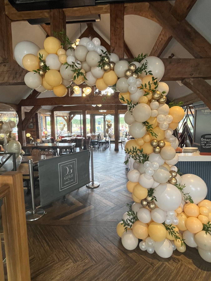 Decorative balloon arch with white, yellow, and gold balloons in a room with wooden beams.