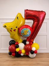 Load image into Gallery viewer, A birthday balloon arrangement featuring a large metallic red number 7 balloon, a yellow Pikachu star-shaped foil balloon, a round Pikachu medallion balloon, and a cluster of red, black, white, and yellow balloons on a wooden floor against a white paneled wall.

