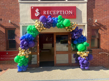 Load image into Gallery viewer, A colorful balloon arch in green, purple, blue, and gold balloons frames the entrance to a brick building reception area with a red sign reading 'RECEPTION'.
