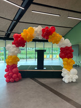 Load image into Gallery viewer, A large balloon arch decoration made of red, yellow, and white balloon clusters, set up in a modern indoor space with glass windows and a black pillar in the background.
