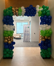 Load image into Gallery viewer, A large balloon arch entrance decoration in blue, green, white, and gold, framing a hallway with a welcome banner visible in the background.

