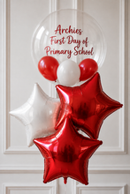 Load image into Gallery viewer, A cluster of party balloons including two metallic red star-shaped foil balloons, one metallic white star-shaped foil balloon, red and white round latex balloons, and a large clear round balloon printed with the text 'Archies First Day of Primary School' in red script, set against a white paneled wall.
