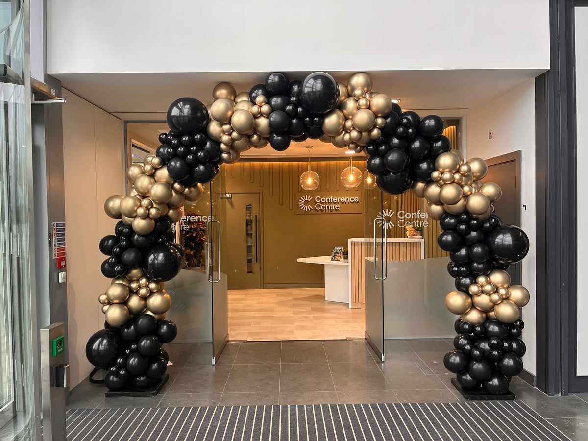 Balloon archway at the entrance of a conference center with black, gold, and silver balloons.